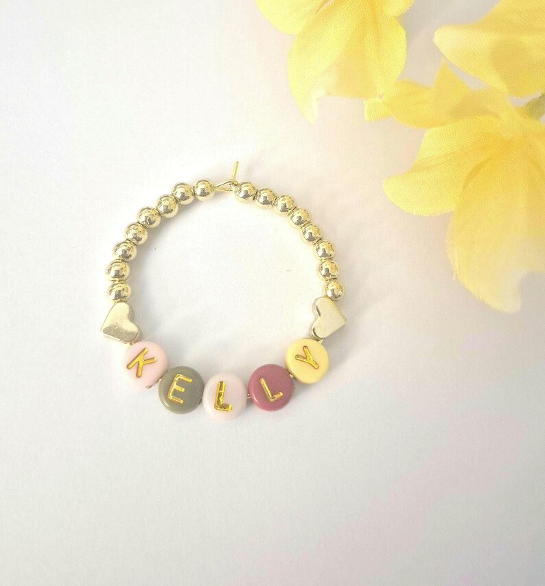 May include: Gold beaded bracelet with the name "KELLY" spelled out in colorful letter beads. The bracelet includes gold heart-shaped charms and is set against a white background with yellow flowers.