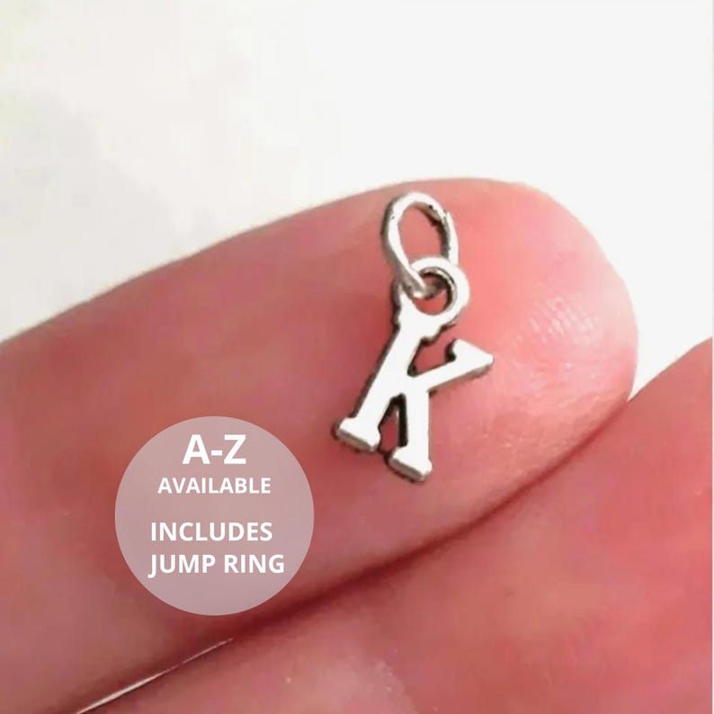 May include: A small silver-toned metal charm in the shape of the uppercase letter K, featuring a circular jump ring attached to the top. The text A-Z AVAILABLE INCLUDES JUMP RING is printed in white on a circular overlay on the image.