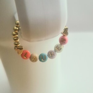 May include: A gold beaded bracelet with the name "LAURE" spelled out in colorful letter beads. The bracelet features gold heart-shaped accents and is displayed on a white surface.