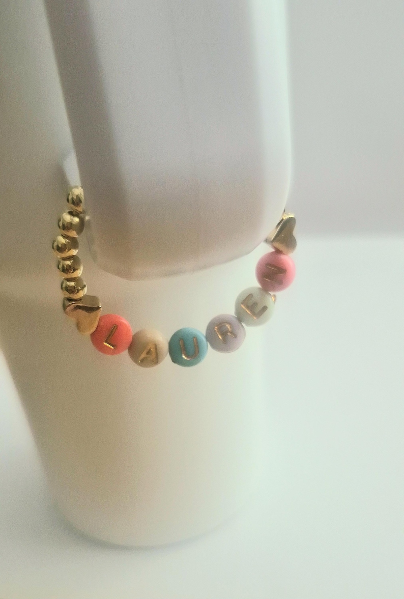 May include: A gold beaded bracelet with the name "LAURE" spelled out in colorful letter beads. The bracelet features gold heart-shaped accents and is displayed on a white surface.