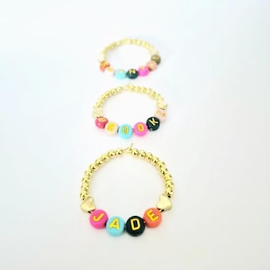May include: Three gold beaded bracelets with colorful letter beads spelling out names. The bracelets have gold heart-shaped accents. The names are JADE, ROOK, and another bracelet with an unreadable name.