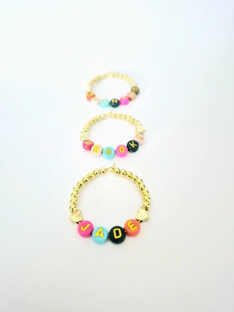 May include: Three gold beaded bracelets with colorful letter beads spelling out names. The bracelets have gold heart-shaped accents. The names are JADE, ROOK, and another bracelet with an unreadable name.