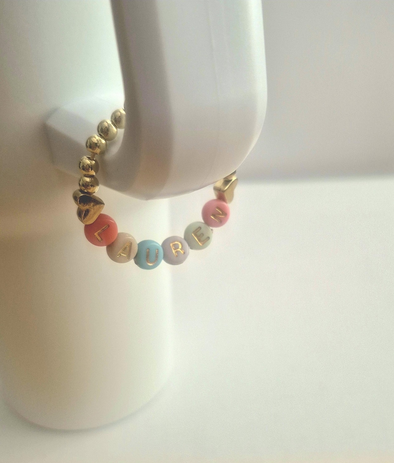 May include: A gold beaded bracelet with the name "LAUREN" spelled out in colorful letter beads. The bracelet is held by a white object. The bracelet has gold heart-shaped accents.
