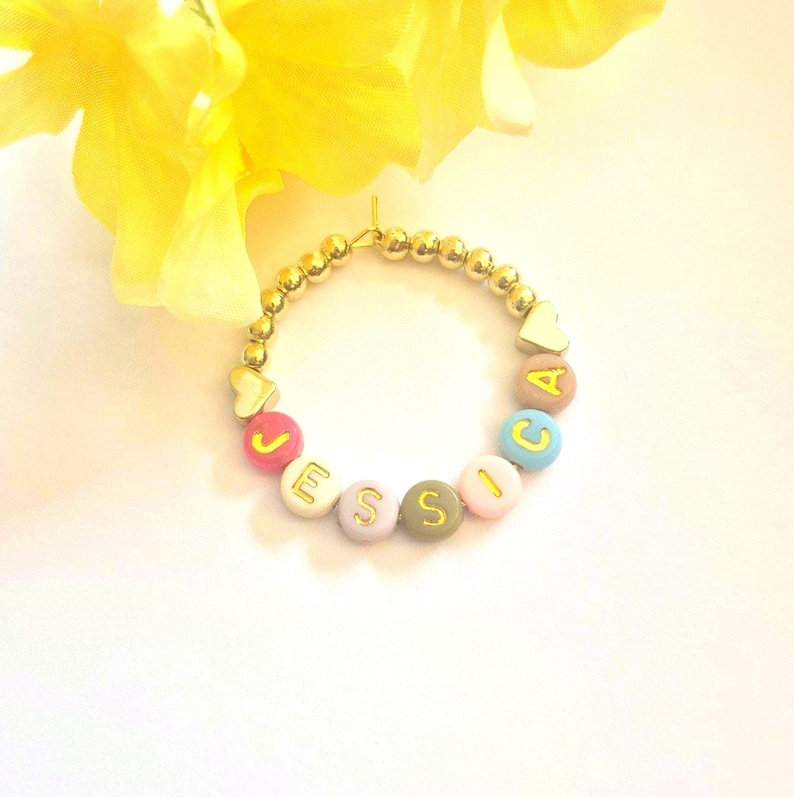 May include: A gold beaded bracelet with colorful letter beads spelling "Jessica". The bracelet features gold heart-shaped charms and is set against a white background with a yellow flower.