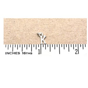 May include: A silver-colored metal charm in the shape of the letter K, featuring a small circular jump ring at the top. The charm is positioned above a ruler showing measurements in inches, indicating a height of approximately 0.25 inches.