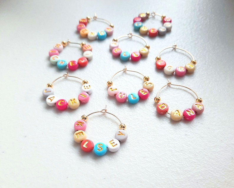 May include: Set of nine gold hoop wine glass charms with colorful beads spelling out names. The names are JACKI, JULES, SEL, DAN, LORI, BLESS,  KELLY, and two others.
