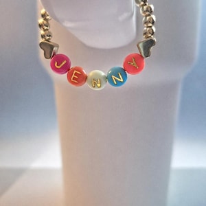May include: A gold-colored beaded bracelet with the name "JENNY" spelled out in colorful letter beads. The bracelet has heart-shaped accents and is displayed against a white background.