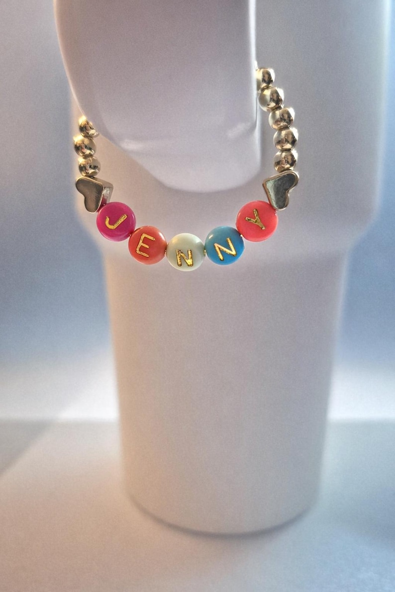 May include: A gold-colored beaded bracelet with the name "JENNY" spelled out in colorful letter beads. The bracelet has heart-shaped accents and is displayed against a white background.