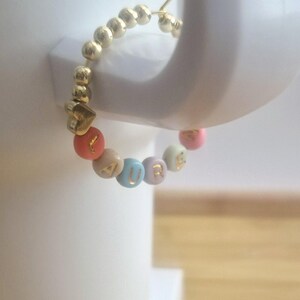 May include: Gold hoop earring with a series of small gold beads. The earring features colorful beads spelling "CURE" with a gold heart charm. The beads are in shades of pink, orange, beige, and blue.