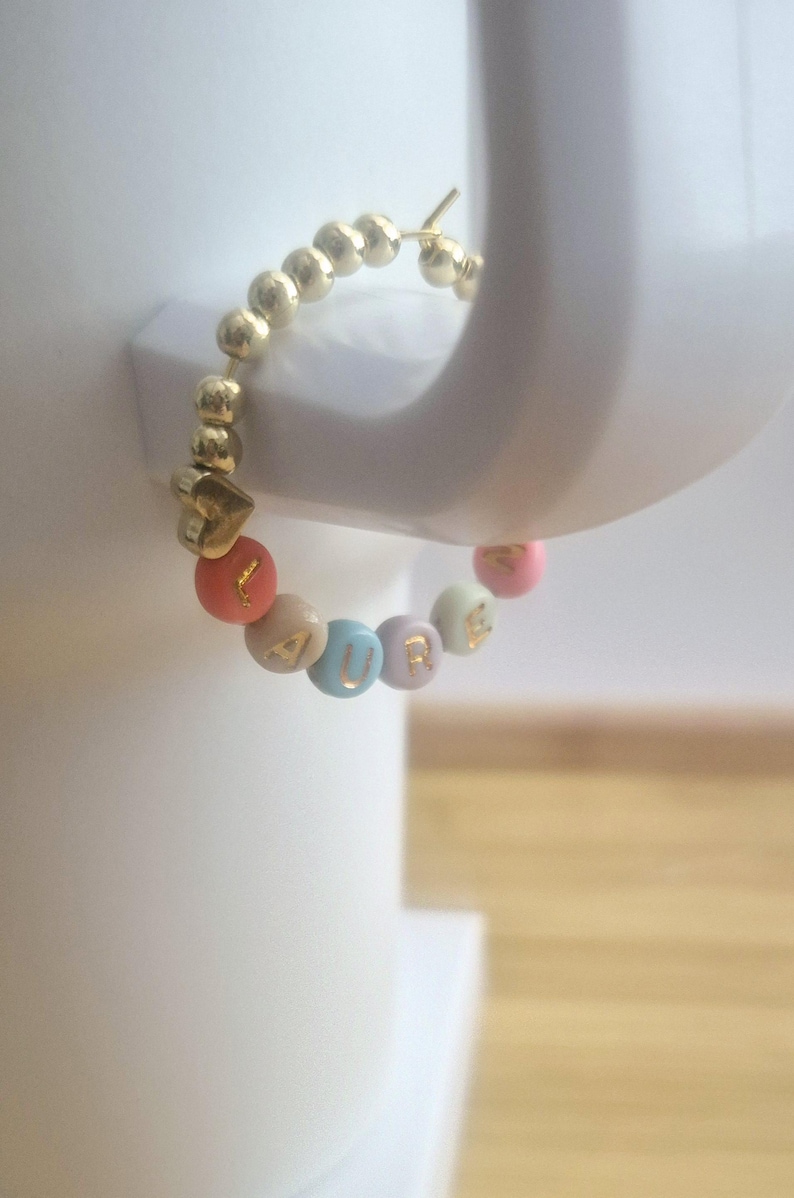 May include: Gold hoop earring with a series of small gold beads. The earring features colorful beads spelling "CURE" with a gold heart charm. The beads are in shades of pink, orange, beige, and blue.