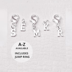 May include: Silver-toned metal letter charms featuring the letters S, E, M, K, and R. Some charms include lobster claw clasps, while others have small jump rings. A circular white graphic displays the text A-Z AVAILABLE INCLUDES JUMP RING.