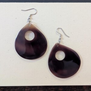 May include: A pair of teardrop-shaped earrings with a dark brown and tan gradient. Each earring has a circular cutout near the center and a silver-colored hook. The earrings are displayed on a light-colored surface.