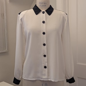 May include: A cream-colored blouse with black accents, displayed on a mannequin. The blouse features a black collar, shoulder details, and cuffs. It has a button-down front with dark buttons. The text "ROYAL FORM" is visible on the mannequin.