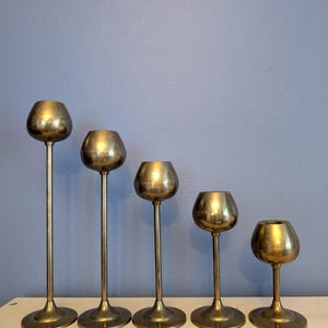 May include: Five brass candle holders of varying heights. Each holder has a round base, a slender stem, and a cup-shaped top. The candle holders are arranged in a row, with the tallest on the left and the shortest on the right.