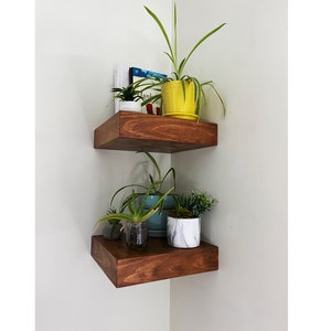 Peut inclure: Deux étagères d'angle en bois avec des plantes en pots. L'étagère du haut a un pot jaune avec une plante verte et un livre. L'étagère du bas a un pot bleu, un pot blanc et une plante verte.