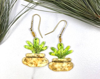 以下が含まれることがあります： 緑色の植物のデザインが施された、ゴールドのドロップイヤリングのペア。植物は、茶色と白の斑点模様の白いセラミックベースに描かれています。