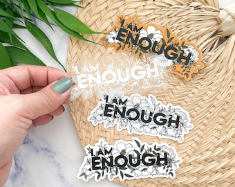 May include: A collection of stickers with the phrase "I AM ENOUGH" in black and white lettering, surrounded by floral designs. One sticker is on a transparent background, and another is on an orange background. The stickers are held by a hand.