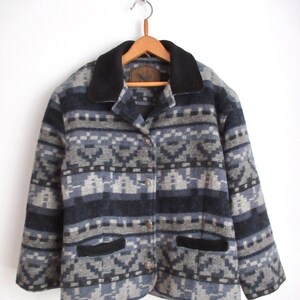 Puede incluir: Una chaqueta con estampado azul, gris y negro con cuello y bolsillos negros. La chaqueta tiene cierre de botones y está hecha de un material suave y peludo.