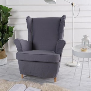 May include: A gray wingback armchair with a high back and a tufted seat. The chair is on a white rug in front of a white wall.