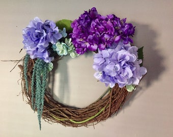 May include: A grapevine wreath decorated with purple, lavender, and light blue hydrangeas.