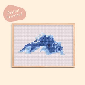 Op de afbeelding: Een ingelijst kruissteekpatroon met een ontwerp van een bergketen in blauw- en lichtblauwtinten. Het kunstwerk staat tegen een witte achtergrond en is omlijst in licht hout. De woorden "Digital Download" staan in een roze bubbel linksboven.