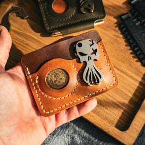 Peut inclure: Gros plan sur deux portefeuilles en cuir avec des détails métalliques, dont un tenu en main. Les portefeuilles sont marron et vert olive, avec des motifs d'arêtes de poisson et de crânes. Autres accessoires : un ouvre-bouteille et un multi-outil.