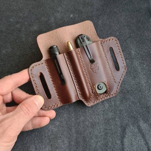 Pode incluir: Um organizador de bolso de couro marrom com três slots para carregar uma caneta, uma lanterna e uma faca dobrável. O organizador tem um design costurado e uma aba com fecho de pressão.
