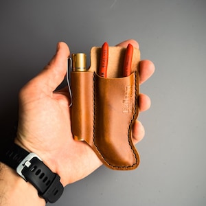 Könnte beinhalten: Ein brauner Leder-Werkzeughalter mit zwei Taschen. Der Halter hat einen Stift oder eine Taschenlampe in Messingfarbe in einer Tasche und eine rote Zange in der anderen Tasche. Der Halter wird in der Hand einer Person gehalten.