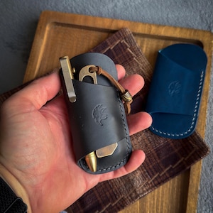Könnte beinhalten: Ein schwarzer Leder-Taschenorganizer mit eingeprägtem Adler-Logo, der einen Messingstift und ein Messingwerkzeug mit brauner Lederschnur enthält. Eine separate blaue Lederhülle mit weißen Nähten und Adler-Prägung liegt im Hintergrund.