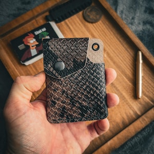 Può includere: Un portafoglio in pelle marrone e nera con un motivo strutturato, una chiusura a scatto e un piccolo occhiello in metallo. Il portafoglio è tenuto in mano e poggia su un vassoio di legno con una moneta, un pettine e una carta.