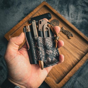 Può includere: Un organizer da tasca in pelle nera con una tasca marrone a motivo di pelle di serpente, contenente una penna, un coltello e altri piccoli strumenti. L'organizer è tenuto in mano, con un vassoio di legno sullo sfondo.