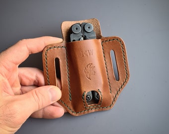 May include: A brown leather multi-tool pouch with green stitching. The pouch has a belt loop and a flap that secures with a snap. The pouch is designed to hold a multi-tool with a pocket knife, pliers, and other tools. The pouch has the initials "KW" embossed on the front.
