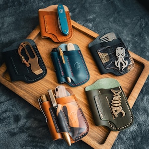 Puede incluir: Una colecci&oacute;n de organizadores de bolsillo de cuero hechos a mano en varios colores, cada uno con un cuchillo peque&ntilde;o, un bol&iacute;grafo o una herramienta. Los organizadores se exhiben en una bandeja de madera, mostrando los diferentes dise&ntilde;os y texturas.
