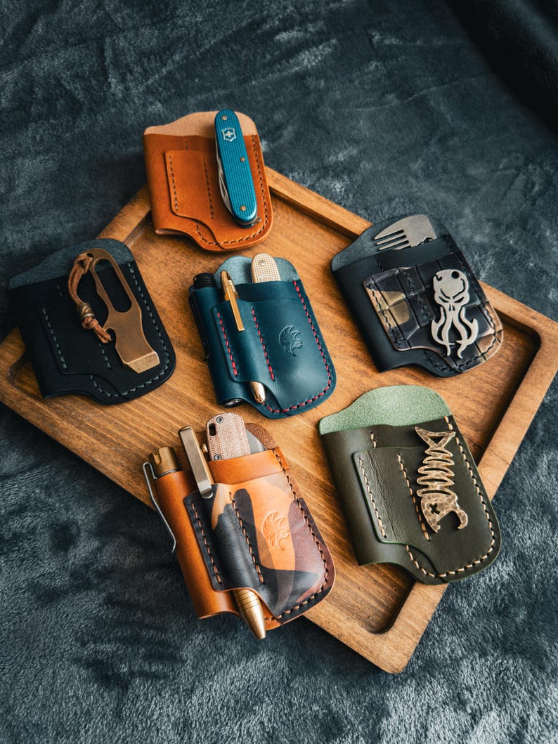 Puede incluir: Una colecci&oacute;n de organizadores de bolsillo de cuero hechos a mano en varios colores, cada uno con un cuchillo peque&ntilde;o, un bol&iacute;grafo o una herramienta. Los organizadores se exhiben en una bandeja de madera, mostrando los diferentes dise&ntilde;os y texturas.