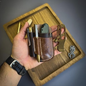 Puede incluir: Un organizador de bolsillo de cuero marr&oacute;n con un bol&iacute;grafo, un cuchillo y otros accesorios. Tambi&eacute;n se ve un abrebotellas de lat&oacute;n y un peque&ntilde;o dije con forma de pulpo. Presentado sobre una bandeja de madera.