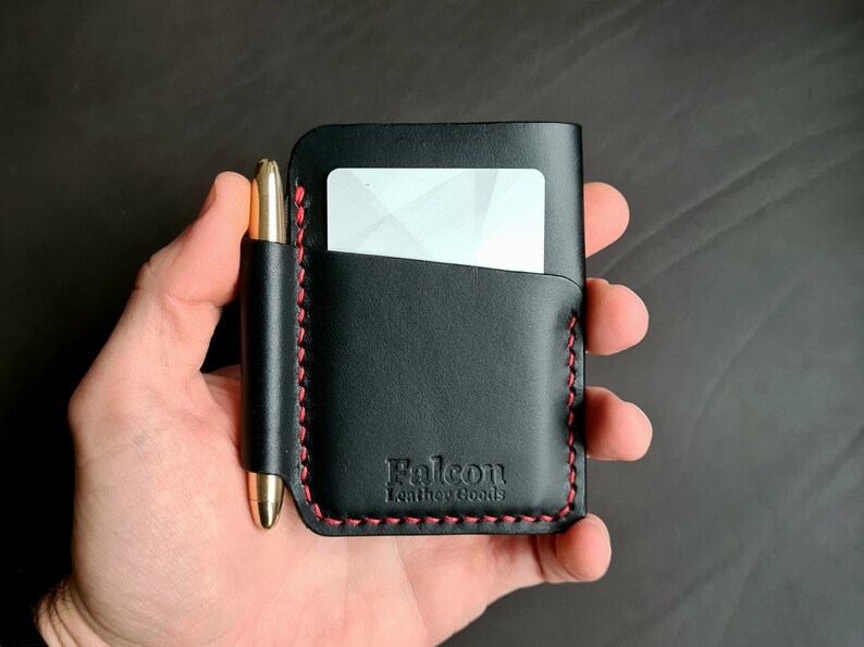 Puede incluir: Cartera de cuero negro con detalles de costura roja y un portaplumas. La cartera tiene una ranura para tarjetas y el texto "Falcon Leather Goods" est&aacute; estampado en la parte delantera.