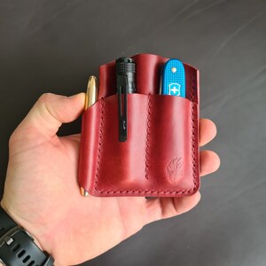 May include: A red leather pocket organizer with three slots. The organizer is holding a pen, a flashlight, and a blue pocket knife.