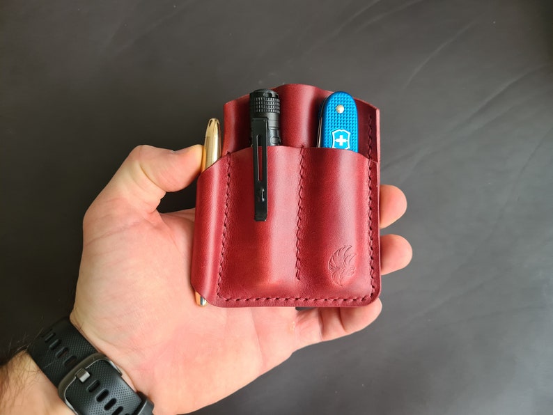 May include: A red leather pocket organizer with three slots. The organizer is holding a pen, a flashlight, and a blue pocket knife.