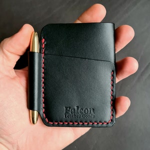 Puede incluir: Cartera de cuero negro con detalles de costura roja y un portaplumas. La cartera tiene el texto "Falcon Leather Goods" grabado en relieve en la parte delantera.