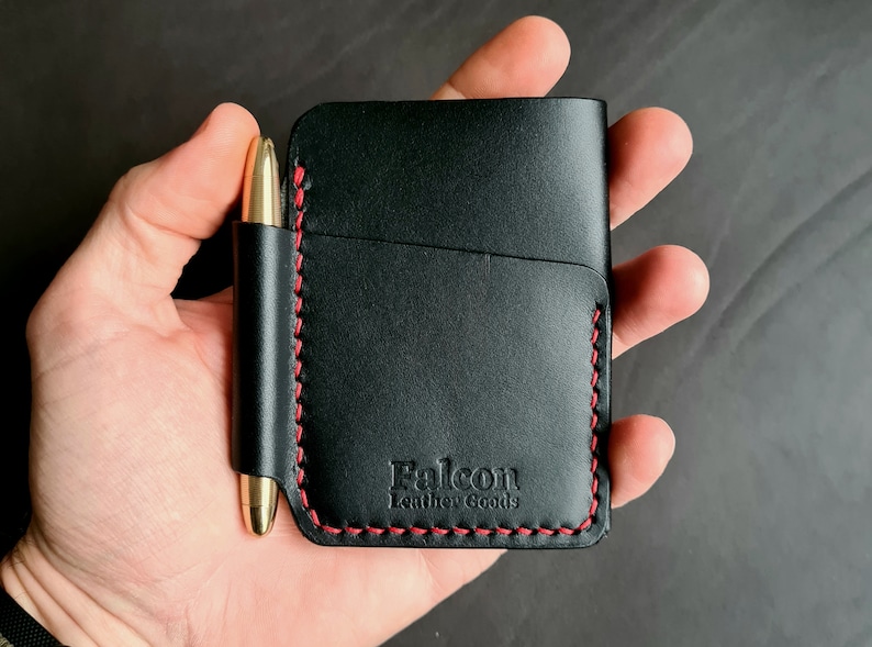 Puede incluir: Cartera de cuero negro con detalles de costura roja y un portaplumas. La cartera tiene el texto "Falcon Leather Goods" grabado en relieve en la parte delantera.