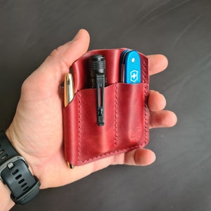 May include: A red leather pocket organizer with three slots. The organizer contains a pen, a flashlight, and a blue pocket knife.