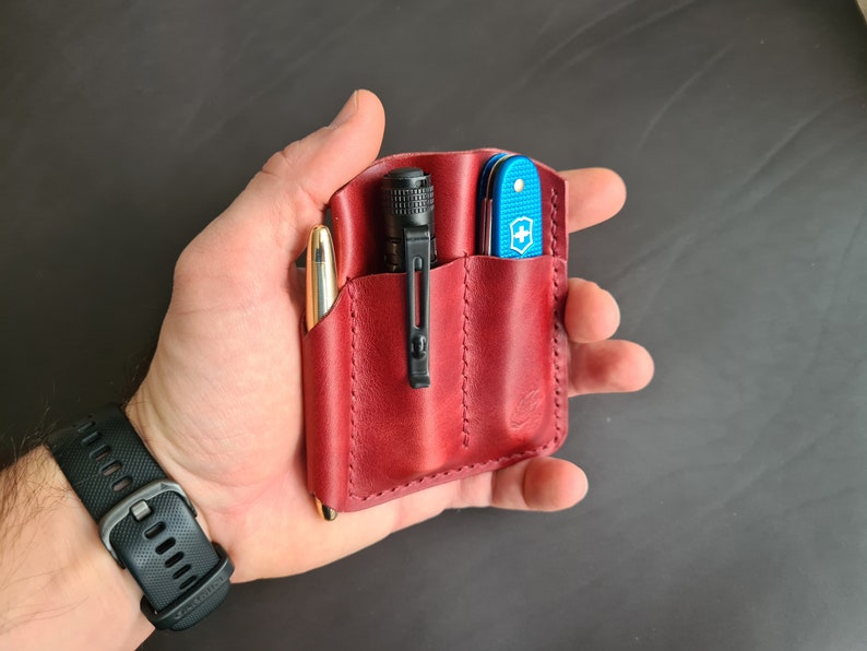 May include: A red leather pocket organizer with three slots. The organizer contains a pen, a flashlight, and a blue pocket knife.