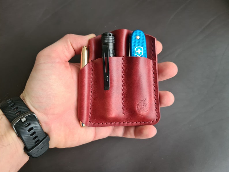 May include: A red leather carrying case with three pockets. The case has a stitched edge and a small logo on the front. The case is holding a black flashlight, a blue pocket knife, and a gold pen.