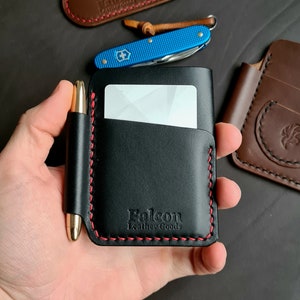Puede incluir: Cartera de cuero negro con costura roja y un portaplumas. La cartera tiene una ranura para tarjetas y el texto "Falcon Leather Goods" est&aacute; estampado en la parte delantera.