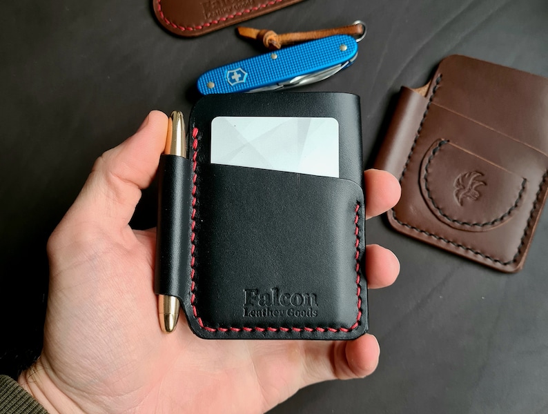 Puede incluir: Cartera de cuero negro con costura roja y un portaplumas. La cartera tiene una ranura para tarjetas y el texto "Falcon Leather Goods" est&aacute; estampado en la parte delantera.