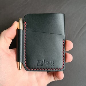 Puede incluir: Cartera de cuero negro con detalles de costura roja y un portaplumas. La cartera tiene el texto "Falcon Leather Goods" grabado en la parte delantera.