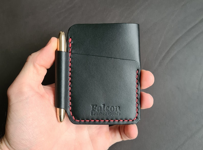 Puede incluir: Cartera de cuero negro con detalles de costura roja y un portaplumas. La cartera tiene el texto "Falcon Leather Goods" grabado en la parte delantera.