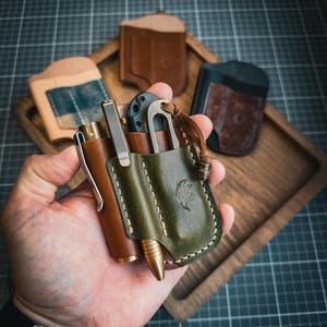 Puede incluir: Una colecci&oacute;n de art&iacute;culos de cuero EDC (Everyday Carry). La imagen presenta un portal&aacute;pices de cuero marr&oacute;n, una funda multiherramienta de cuero verde y varias carteras de cuero en una bandeja de madera. El portal&aacute;pices y la funda se sostienen en una mano.