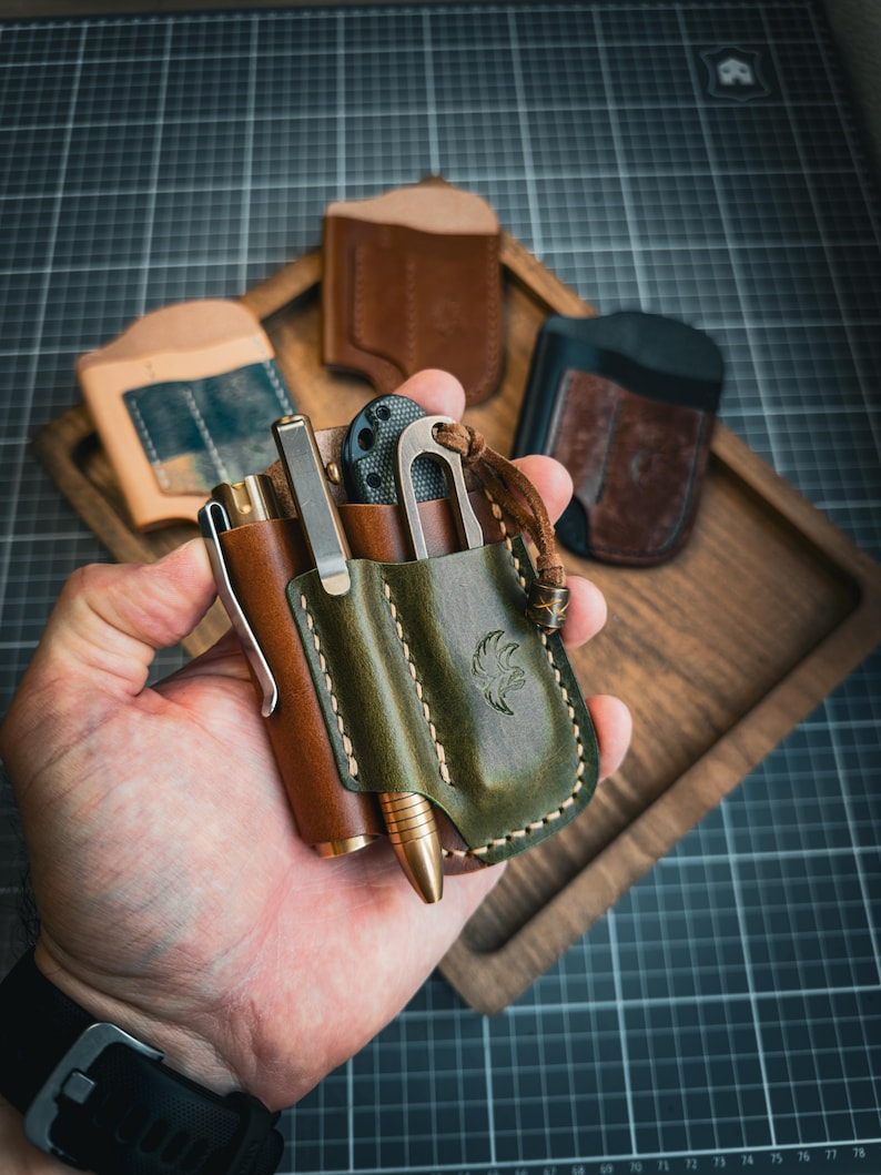 Puede incluir: Una colecci&oacute;n de art&iacute;culos de cuero EDC (Everyday Carry). La imagen presenta un portal&aacute;pices de cuero marr&oacute;n, una funda multiherramienta de cuero verde y varias carteras de cuero en una bandeja de madera. El portal&aacute;pices y la funda se sostienen en una mano.