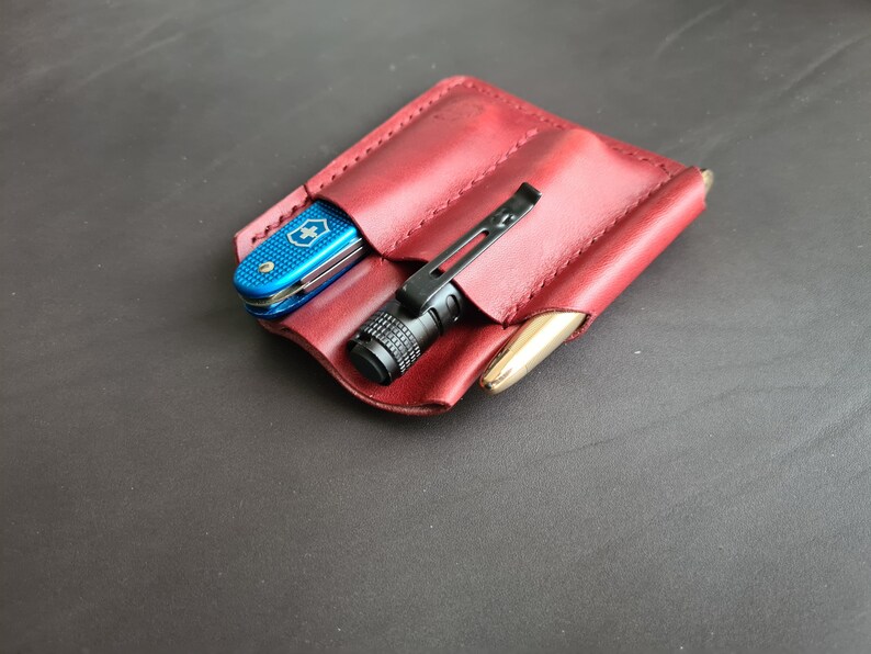 May include: A burgundy leather pocket organizer with three slots. A blue pocket knife, a black flashlight, and a gold pen are in the slots.